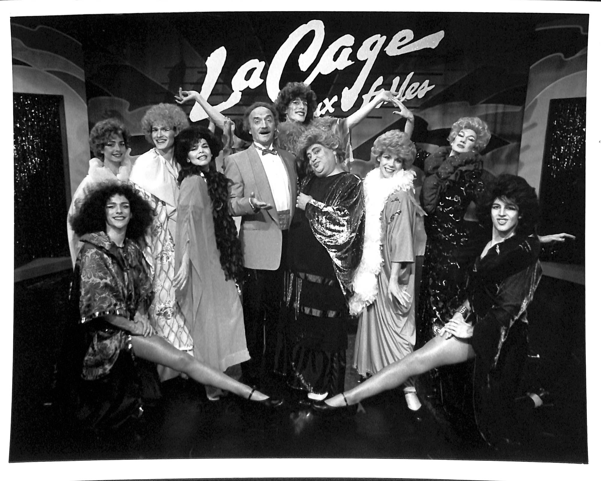A black and white photo of nine women and a man who pose in front of a set that reads "La Cage." They are all wearing costmes and two are seated with their toes pointed in front of the others. The back of "La Cage," which lists the cast from left to right.